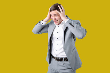 A man in a gray jacket and white shirt on a bright yellow background. He holds his head, expressing surprise or fear. His posture and facial expression convey strong emotions.