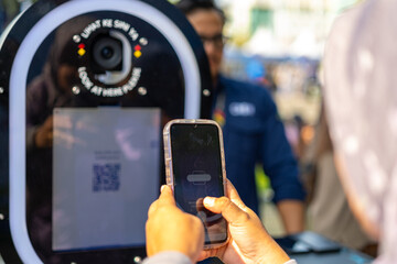 Close-up of a person using a smartphone to interact with a modern photo booth featuring a camera lens and QR code display during an outdoor public event.