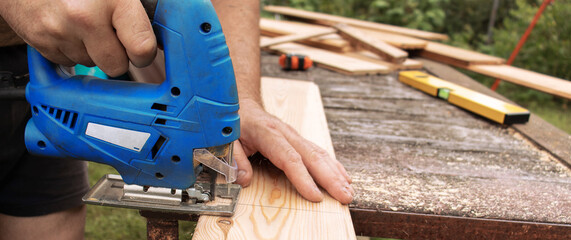 Close up of male hands of unknown carpenter working with electric jigsaw cutting wood with saw,...