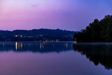 lakeside scene with early morning sunrise colour reflecting with distant lights in a still calm lake silhouetting trees, Lake Windermere, The Lake District, Cumbria, England