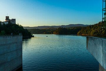 Obraz premium A view of Czorsztyn Lake with Niedzica Castle and distant hills under soft evening light. A boat drifts quietly across the water, emphasizing peace, tranquility, and the harmony of history with nature