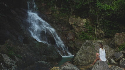 Woman sit on stone enjoy jungle mountain waterfall. Female relax looking on splashing water. Girl in white shirt enjoy beautiful wild nature landscape, exotic rainforest. Dark toning