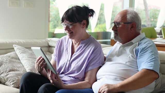 Elderly man showing woman how to use a tablet while sitting on a couch, smiling and enjoying digital learning together in a modern living room.
