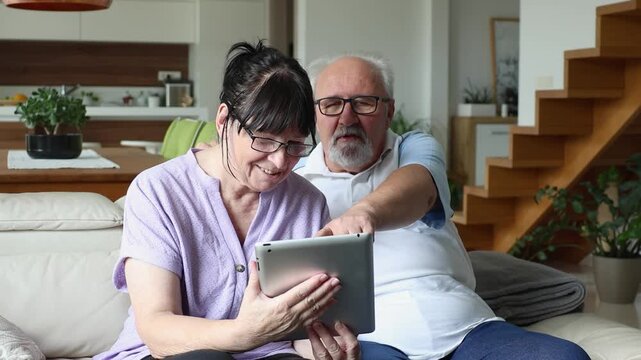 Elderly man and woman sitting on a couch at home, smiling together while looking at a digital tablet, enjoying technology and leisure.