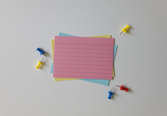Top view of blank index cards and push pins on a white background. A stack of colorful empty note cards and pins. Note-taking, organization, and office supplies concepts.