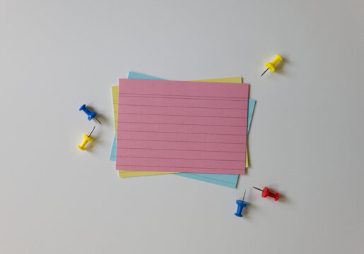 Top view of blank index cards and push pins on a white background. A stack of colorful empty note cards and pins. Note-taking, organization, and office supplies concepts.