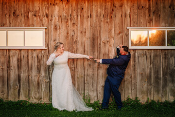 Bride and groom in wedding attire strike a playful pose pointing at each other in front of a rustic wooden wall during golden hour, creating a fun wedding portrait. © Raivo