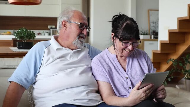 lderly man and woman sitting on a couch, smiling while looking at a tablet and learning technology together in a modern home environment.