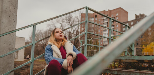 A young woman in a cozy jacket sits on outdoor steps surrounded by autumn leaves in an urban...