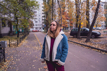 A woman stands thoughtfully in an urban setting during autumn, surrounded by trees and parked cars...