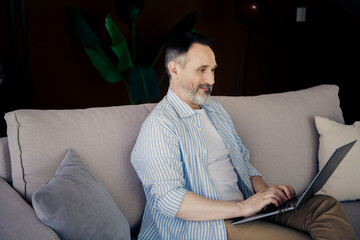Smiling mature man working remotely on laptop from comfortable modern living room suggesting...