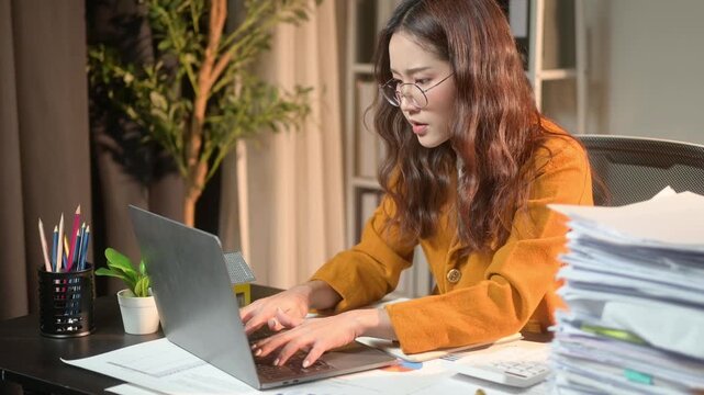 woman is sitting at a desk with a laptop and stacks of papers. She is typing on the laptop and she is focused on her work. Concept of productivity and concentration as the woman works on her tasks