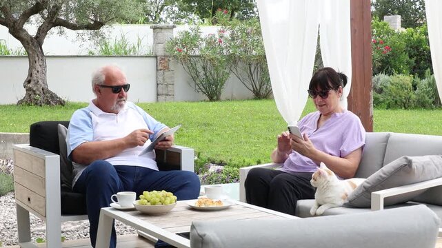 Elderly man and woman sitting on a garden patio with their cat, relaxing while using smartphone and tablet, enjoying leisure time and technology.