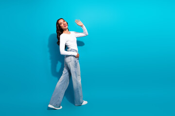 Young cheerful woman with long brunette hair waving on vibrant blue background, showcasing casual...