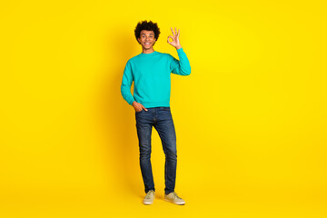 Cheerful young man in stylish teal sweatshirt showing ok hand gesture against bright yellow background in casual style