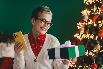 Cheerful mature woman holding a festive gift and smartphone by the Christmas tree in a cozy holiday...