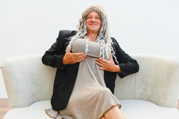 Man wearing wig and dress showing fake breasts while sitting on sofa