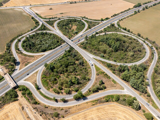Aerial view of a symmetrical cloverleaf highway interchange. Cars on the road network show transportation and logistics in a rural landscape, symbolizing connectivity and infrastructure.