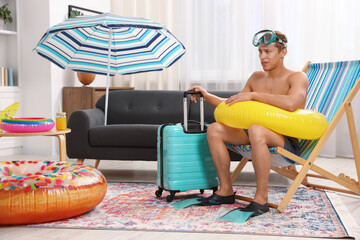 Man in flippers with diving mask, suitcase and inflatable ring on folding chair at home. Dreaming...