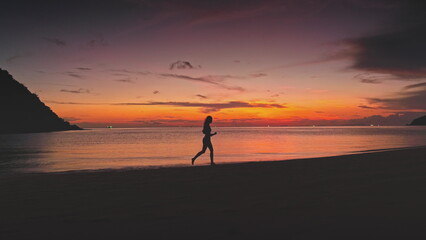 Silhouette of an athletic woman running along the ocean coast at sunset, with a vibrant sky filled with colorful clouds reflecting on the tranquil water surface in Thailand