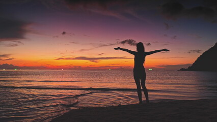 Fototapeta premium Silhouette of young woman opening arms, enjoying freedom and idyllic colorful sunset at tropical beach in Thailand, with calm sea water and some boats in the distance
