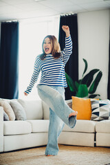 Joyful woman enjoying leisure time and dancing in living room wearing striped casual outfit
