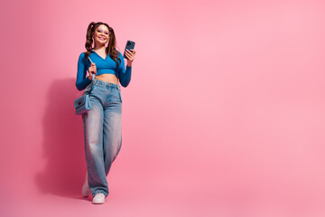 Young stylish woman with ponytail hairstyle wearing casual blue top using smartphone while posing against pink wall