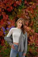 Young woman in casual autumn outfit posing against vibrant red ivy wall, stylish fashion portrait in warm seasonal tones.