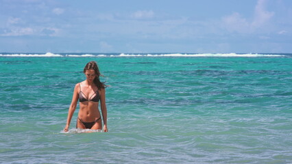 A woman wearing a bikini is standing in the ocean water. The sun is shining down on her as waves crash around her. She is enjoying the beach and the water.