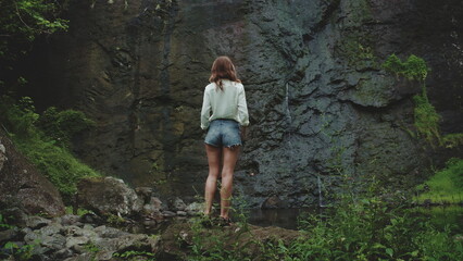Naklejka premium Tourist is standing on a rock, admiring a waterfall flowing on a volcanic rock wall. The lush vegetation surrounding the area creates a sense of tranquility and adventure