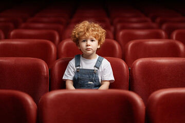 Obraz premium A young boy sits alone in an empty theater, gazing at the audience with a thoughtful expression. The red seats add a dramatic contrast to his appearance.