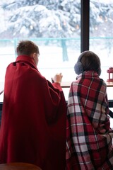 Two individuals bundled in warm blankets, gazing out a window at a snowy winter scene.