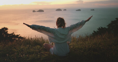 Woman sit grassy hilltop with arms outstretched, meet sunrise over ocean. She enjoy peaceful scenery and warm glow of morning light. Freedom and tranquility, beauty of Thailand tropical island nature
