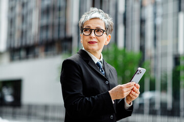Businesswoman standing outdoors urban setting holding mobile phone in elegant formal jacket and...
