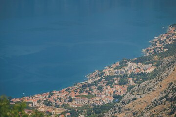 Coastal houses and buildings nestle along the shoreline of Kotor Bay, under a bright, sunny sky....