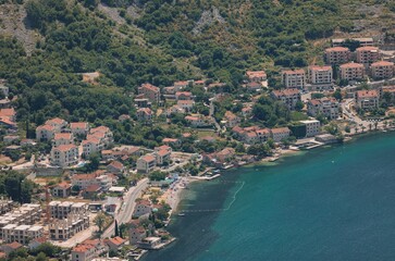 Naklejka premium Along the Adriatic coast, buildings cluster the shore, climbing the mountainside. People relax on a narrow beach beside turquoise water in the summer sunshine near Kotor