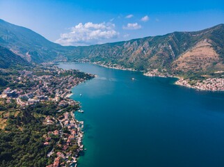 Obraz premium A deep blue bay is nestled between lush green mountains and charming coastal towns in Montenegro. Small boats navigate the tranquil waters under a bright blue sky