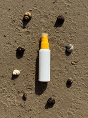 A white plastic spray bottle with a yellow cap placed on wet sand, surrounded by seashells. Minimalist beach-themed composition, suitable for concepts of summer, skincare, sunscreen, vacation