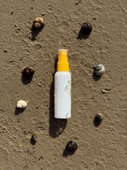 A white plastic spray bottle with a yellow cap placed on wet sand, surrounded by seashells. Minimalist beach-themed composition, suitable for concepts of summer, skincare, sunscreen, vacation