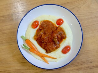 Mashed potatoes topped with tomato meat sauce, cherry tomatoes, and carrots on white-blue plate. Taken at casual restaurant in Bangkok, Thailand.