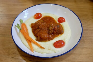 Mashed potatoes topped with tomato meat sauce, cherry tomatoes, and carrots on white-blue plate. Taken at casual restaurant in Bangkok, Thailand.