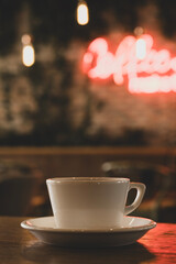 Modern cafe concept with coffee cup and neon glow. White coffee cup with neon sign background. Hot coffee in front of blurred neon lights. Trendy coffee cup with vibrant neon background.