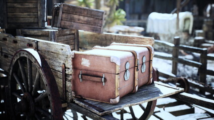 A rustic wooden cart holds two weathered leather trunks, set against a backdrop of an old western village under bright sunlight. The scene evokes a sense of nostalgia.