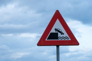 Red triangular warning sign showing car falling into water under cloudy sky