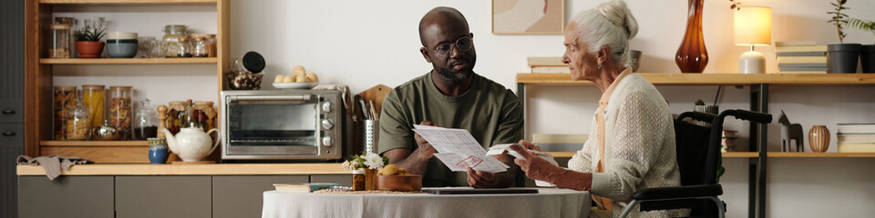 Young Black man explaining documents to senior Caucasian woman with disability sitting in...