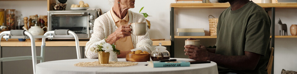 Senior Caucasian woman with disability sitting at table holding mug, talking with young adult Black man in kitchen, both engaging in conversation with coffee and snacks