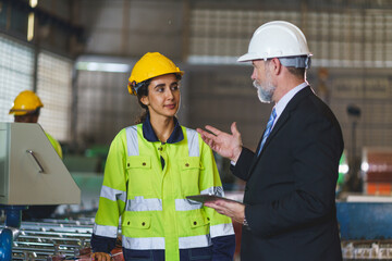A foreman and a woman worker are standing in a factory, looking at a tablet. The engineer talks with the woman's workers. They wear a yellow and white safety helmet for safety.
