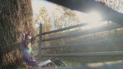 Young woman is relaxing in the sun, sitting by a haystack and reading a book in the peace of the countryside on a beautiful autumn day