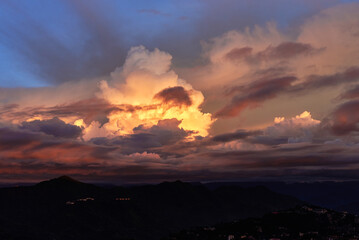 Golden sunset clouds blaze through the evening sky, casting a warm glow over the dark, quiet mountain silhouettes below.
