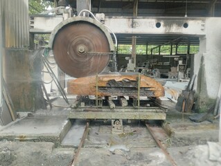 An outdoor shot of an industrial diamond saw cutting through a massive, rough block of natural stone at a workshop. The machine is actively in use, with water spraying to cool the blade & reduce dust